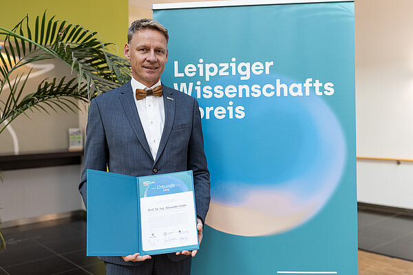 Mann mit Urkunde steht vor Plakat