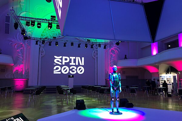 leerer Saal, bunt beleuchtet, mit Androidin auf Bühne in der Mitte